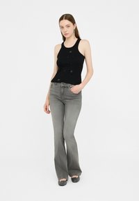 Top nero senza maniche con decorazioni in perline, abbinato a jeans svasati grigi. Il modello indossa scarpe piatte nere, in piedi contro uno sfondo bianco.