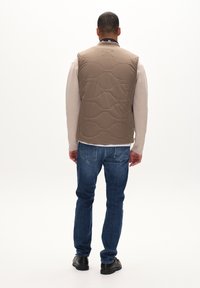 Ljusbrun, kviltad ärmlös väst gjord av mjukt tyg, kombinerad med en beigefärgad långärmad skjorta och blå jeans, vy från baksidan.