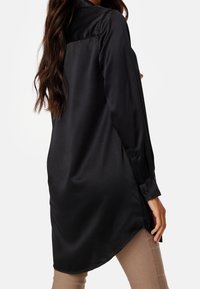 Camisa de satén negra con puños abotonados, mangas largas y dobladillo curvado. Textura suave y diseño de cuello clásico.