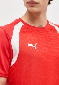 Κοντινό πλάνο ενός ατόμου που φοράει αθλητική μπλούζα Puma κόκκινου και άσπρου χρώματος με διακριτικό σχέδιο που μοιάζει με ιστό σε όλο το ύφασμα.