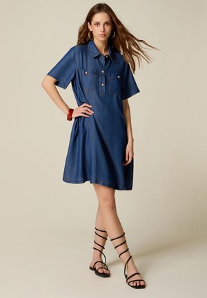 Jonge vrouw met lang haar die een blauwe denim korte mouwen jurk draagt, oorringen, rode armband en zwarte sandalen met bandjes staat met hand op heup.