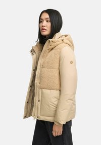 Beige pufferjack met een tan fleecepaneel op de borst, uitgerust met een capuchon, drukknoopsluitingen en zijzakken. Contrasterende textuur van de stof.