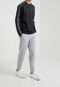 Felpa nera con dettaglio del logo sulle maniche, tasca con zip, abbinata a pantaloni della tuta grigi chiari e sneakers bianche, su uno sfondo neutro.