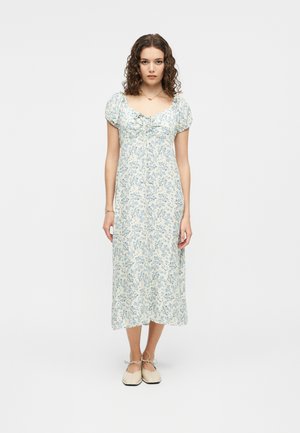 Νεαρή γυναίκα με σγουρά μαλλιά που φοράει λευκό μίντι φόρεμα με μπλε floral σχέδιο και κρεμ μπαλαρίνες, στέκεται μπροστά από ένα απλό φόντο.