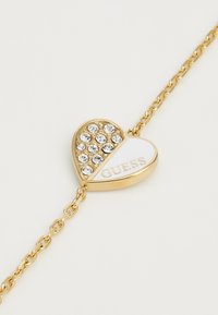 Kuldset tooni südamekujuline käevõru, millel ühe poole peal on selged rhinestone'id ja teisel pool valge email, millel on sõna "GUESS", ühendatud ketiga.
