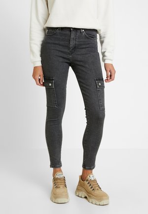 Tight black cargo jeans with flap pockets paired with beige chunky lace-up sneakers and a white long-sleeve top.