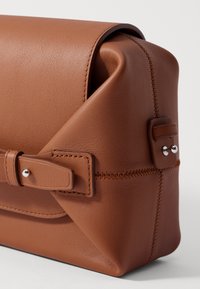 Brown leather handbag with a structured design, visible stitching, and a bow accent, featuring a smooth texture and rounded edges.