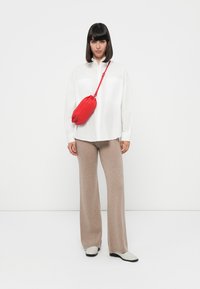 Camicia bianca a maniche lunghe, pantaloni beige a gamba larga e una borsa a tracolla rossa. L'outfit è abbinato a scarpe di colore chiaro. Texture lisce e tonalità neutre.