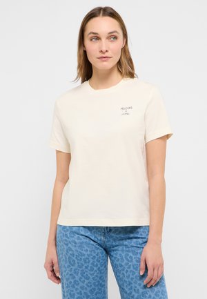 Woman wearing a cream short-sleeve T-shirt with "Mustang Jeans" text and blue leopard print jeans, standing against a plain background.