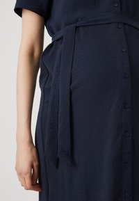 Μπλέ ναυτικού φόρεμα από υφασμάτινη υφή, που διαθέτει κουμπιά κατά μήκος του μπροστινού μέρους και ζώνη δέσμευσης στη μέση για προσαρμοσμένη εφαρμογή.