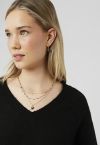 Colliers à couches plaqués or avec des perles noires et transparentes, comportant un pendentif circulaire. Le modèle porte un pull noir à col en V et des boucles d'oreilles assorties.