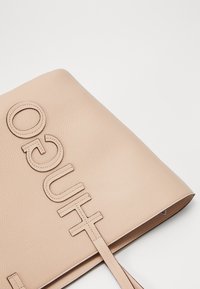 Beige leren tote bag met een geëtst "HUGO" logo, plat ontwerp, twee handvatten en een gladde, gestructureerde oppervlakte.