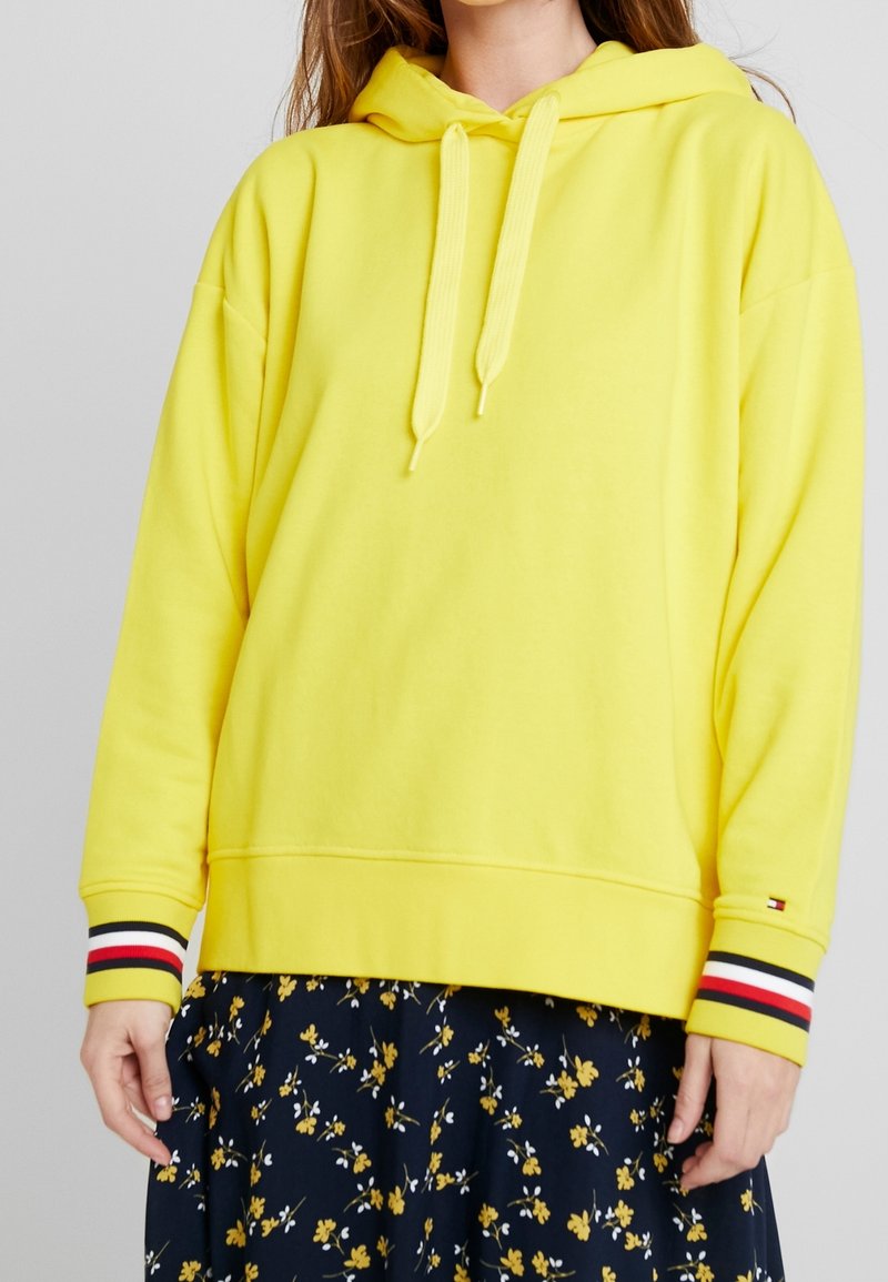 Femme portant un sweat à capuche jaune vif avec des poignets rayés et une jupe noire présentant de petits motifs floraux jaunes et blancs.