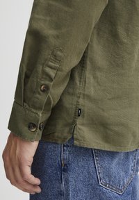 Olijfgroene shirt van een gestructureerde, katoenen stof. Met knoopmanchetten en een zijschuif. Gedragen met blauwe spijkerbroeken.
