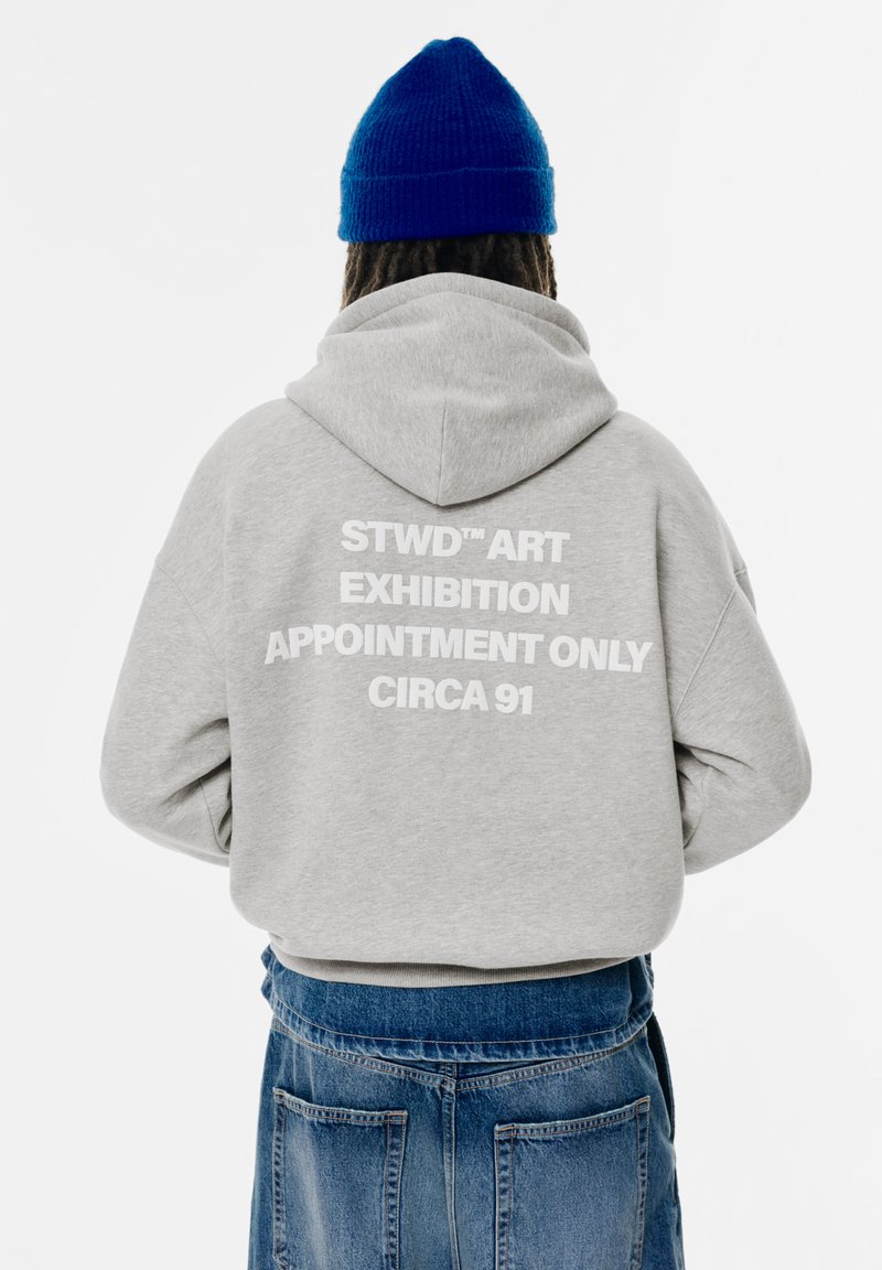 Grå huvtröja med vit text: "STWD™ KONSTUTSTÄLLNING ENDAST EFTER ÖVERENSKOMMELSE CIRKA 91" på baksidan. Stickad blå mössa synlig.