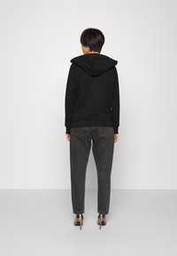 Sweatshirt preto com capuz e corte descontraído, combinado com jeans cinza escuro e saltos brilhantes, visto por trás.