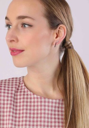 Zilveren haakoorbellen met een dun, verticale stangdesign. Het onderwerp draagt een roze ruitjestop en toont een gladde huid en steil haar.