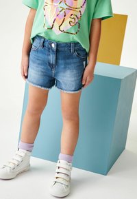 Pantalones cortos de mezclilla con dobladillo deshilachado, lavado azul claro, cinco bolsillos, combinados con una camiseta gráfica verde y zapatillas blancas con cierres de correa.