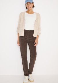 Bruine corduroy broek met een rechte snit, gecombineerd met een crèmekleurige gebreide cardigan en een wit shirt, afgemaakt met lichte sneakers en een blauwe pet.