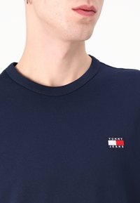 Mann trägt ein dunkelblaues T-Shirt mit Rundhalsausschnitt und kleinem Tommy Jeans Logo auf der linken Brust vor weißem Hintergrund.
