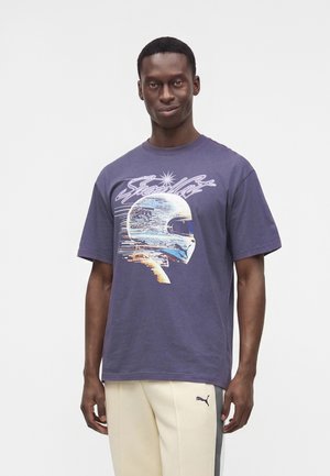 Homme portant un t-shirt violet avec un graphique d'un casque de course blanc et un texte, associé à un pantalon beige et gris Puma.