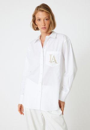 Camisa blanca de botones con collar, que presenta un bolsillo en el pecho izquierdo bordado con "LA" en dorado. Mangas largas, corte relajado, textura suave.