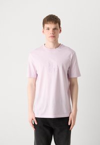 Lyse pink bomulds t-shirt med korte ærmer og rund hals, med et broderet "HUGO" logo på forsiden. Et par sorte jeans.