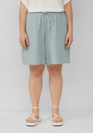 Shorts en coton bleu clair, texturés, avec une taille élastique et un cordon de serrage, portés avec des sandales blanches à lanières, affichant une coupe décontractée.
