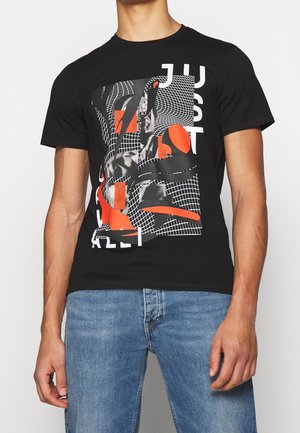 Mężczyzna w czarnej koszulce z abstrakcyjnym pomarańczowym, białym i szarym geometrycznym wzorem oraz częściowym napisem, w połączeniu z niebieskimi dżinsami.