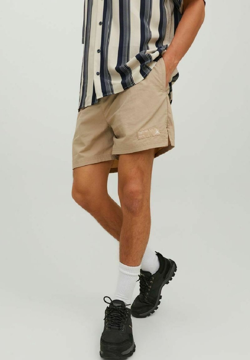 Homme portant un short beige, des baskets noires, des chaussettes blanches et une chemise à manches courtes rayée verticalement noir et blanc, debout avec une main dans la poche.