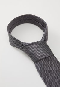 Une cravate grise texturée présente un motif tissé, une extrémité pointue et une finition lisse, adaptée aux occasions formelles.