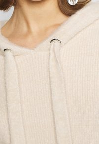 Kremowa bluza z kapturem wykonana z teksturowanej tkaniny, z prążkowanym szwem i srebrnymi oczkami na sznurki.
