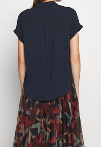 Blouse marine à manches courtes avec col et pli au dos, associée à une jupe plissée à motifs dans des teintes de rouge, marron et vert.