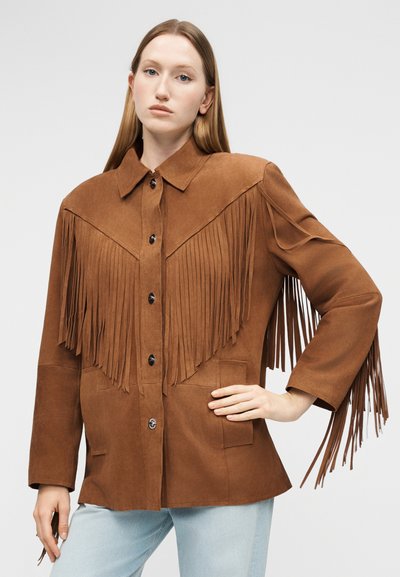 Νεαρή γυναίκα με ίσια μακριά μαλλιά, φοράει καφέ σατέν σακάκι με μακριά κρόσσια στα μανίκια και στο στήθος, συνδυασμένο με ανοιχτόχρωμο μπλε τζιν.