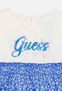 Λευκό τοπ με μπλε γκλίτερ κείμενο "Guess", μπλε φλοράλ φούστα με ελαστικές λεπτομέρειες, κοντά μανίκια και άνοιγμα στην πλάτη με σκίατρο.