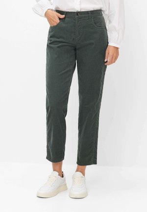 Frau trägt dunkelgrüne Hose mit hohem Bund und geradem Bein, weiße Sneakers und ein weißes langärmeliges Hemd, eine Hand in der Hosentasche.