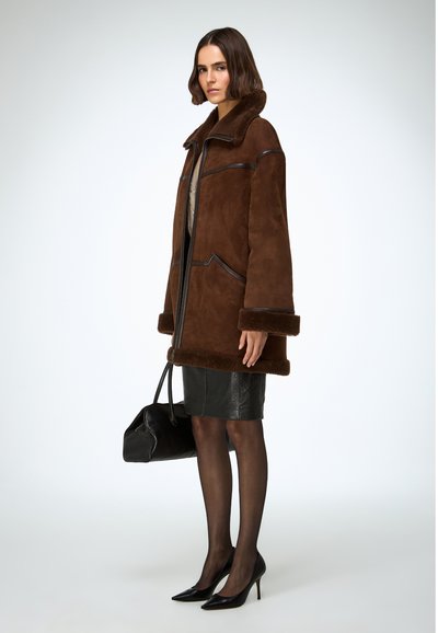 Bruine shearlingjas met een brede kraag, zwarte leren accenten en zachte manchetten, gecombineerd met een zwarte leren rok en puntige hakken.