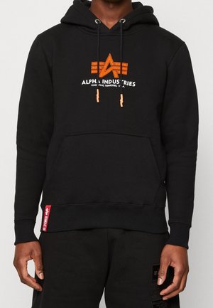 Mężczyzna noszący czarną bluzę z kapturem Alpha Industries z pomarańczowym logo i napisem, przednią kieszenią oraz sznurkami zakończonymi pomarańczowymi końcówkami.