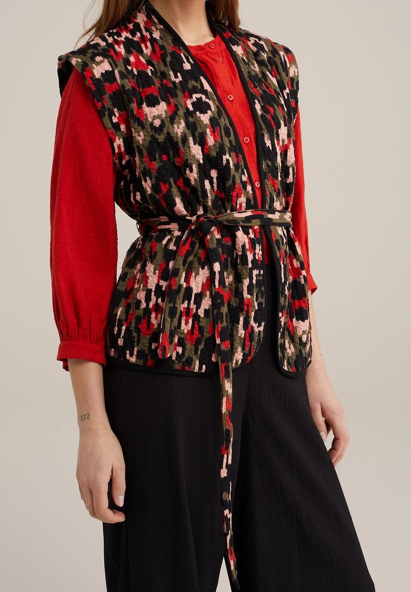 Femme portant un chemisier rouge à manches longues sous un gilet sans manches ceinturé avec un motif abstrait rouge, noir, rose et vert, assorti à un pantalon noir.