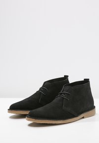 Bottines en daim noir avec un bout pointu, design classique à lacets et semelle en caoutchouc brun clair. Texture lisse et style minimaliste.