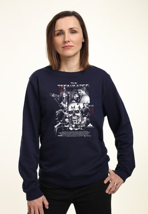Kvinde med skulderlangt brunt hår bærer en navyblå sweatshirt med The Terminator-filmens design med figurer og en robotskalle.