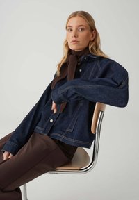 Dunkle Jeansjacke mit großen Taschen und sichtbaren Steppnähten, kombiniert mit einem braunen Strickschal, sitzend auf einem Metallstuhl.