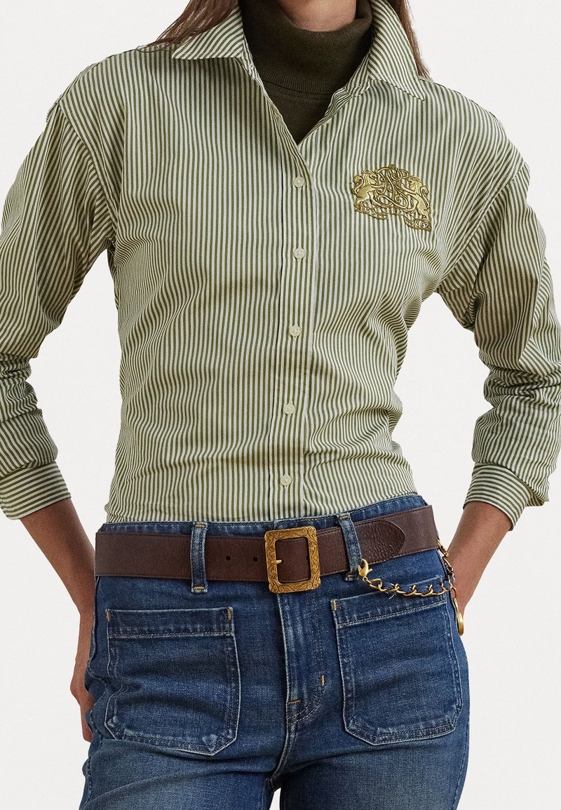 Personne portant une chemise à rayures vertes et blanches à boutons avec un motif brodé doré, un col roulé foncé, un jean bleu et une ceinture marron avec boucle dorée.