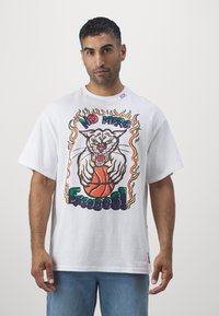 Biela prešívaná tričko s živou grafikou, na ktorej je divoká mačka držiaca basketbal v obklopení plameňmi a textom v farebných písmach.