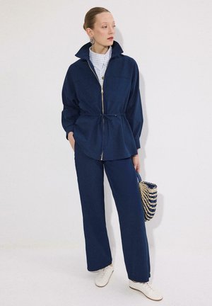 Γυναίκα που φοράει σκούρο μπλε μπουφάν με φερμουάρ και φαρδιά παντελόνια, κρατώντας ριγωτή υφαντή τσάντα, στέκεται μπροστά σε απλό λευκό φόντο.