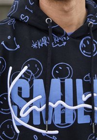 Černá mikina s kapucí, na které jsou modré vzory úsměvů a velká modrá písmena tvořící slovo "SMILE", částečně zakrytá bílým vyšívaným kurzívním textem.