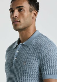 Lichtblauwe gebreide poloshirt met een ruitpatroon, drie knoopsluitingen en geborduurd logo op de borst. Zachte textuur en met kraag ontwerp.