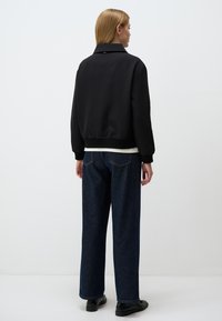 Veste bomber noire avec un col et des poignets côtelés, associée à un jean bleu clair à jambe large. Tissu lisse avec un design minimaliste.