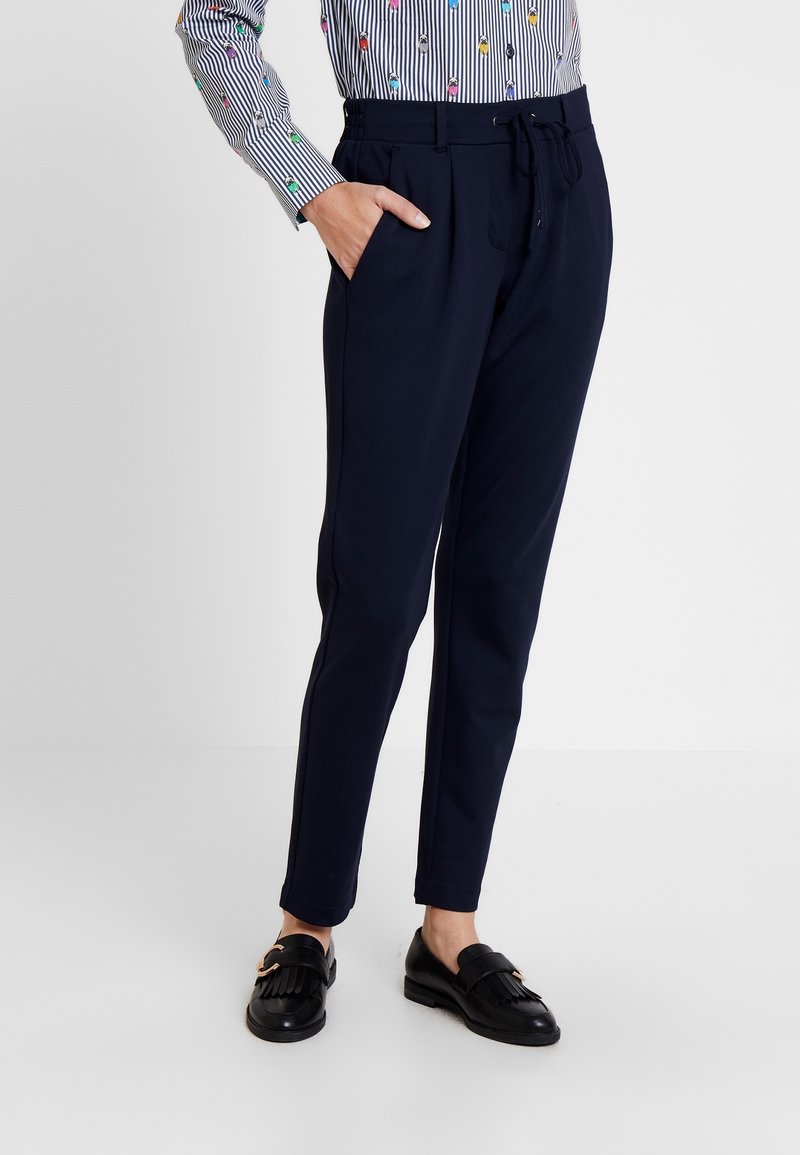 Person som bär marindräktade tapered byxor med sidofickor, svarta loafers med fransdetalj och en randig långärmad skjorta med färgglada broderade mönster.