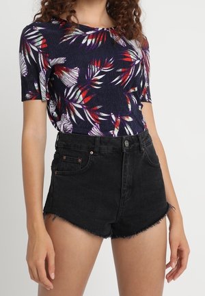 Kobieta w granatowej bluzce z krótkim rękawem z czerwonymi i białymi wzorami liści oraz czarnych, wysokich, dżinsowych szortach z postrzępionymi krawędziami.